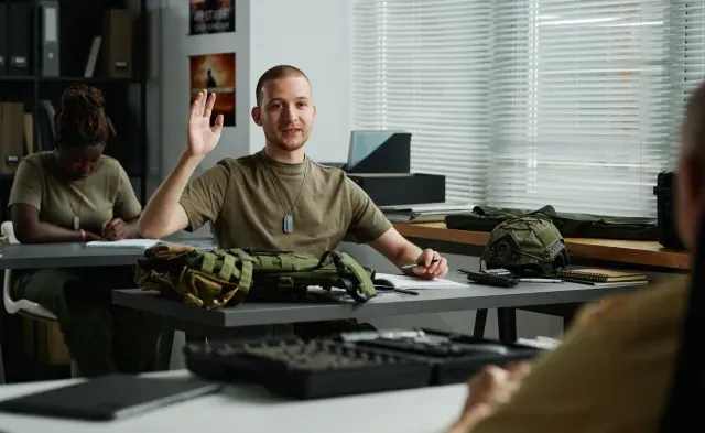 Young male student raising hand and looking at teacher at lesson while asking him question after lecture about military training